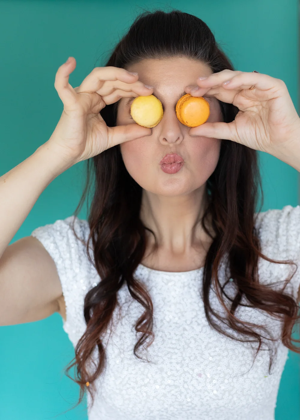 Erica playfully holding two macarons over her eyes against a teal backdrop